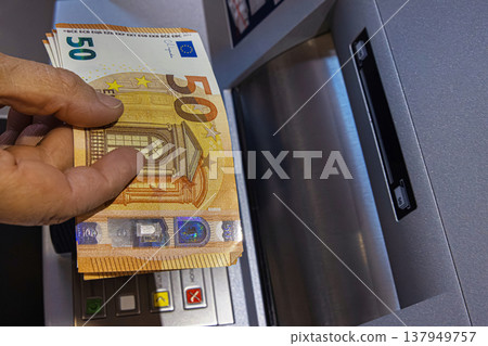 Closeup of a person holding multiple 50 euro banknotes in front of an ATM keypad. The terminal features colored confirmation buttons and a money slot. Closeup of a person holding multiple 50 euro banknotes in front of an ATM keypad. The terminal features colored confirmation buttons and a money slot. 137949757