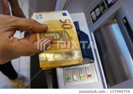 Closeup of a person holding multiple 50 euro banknotes in front of an ATM keypad. The terminal features colored confirmation buttons and a money slot. Closeup of a person holding multiple 50 euro banknotes in front of an ATM keypad. The terminal features colored confirmation buttons and a money slot. 137949758
