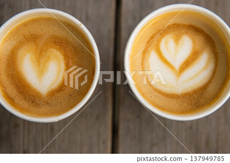 Top down closeup view of two paper coffee cups, placed on a rustic wooden table, with a creamy heart and flower shaped latte art Top down closeup view of two paper coffee cups, placed on a rustic wooden table, with a creamy heart and flower shaped latte art 137949785