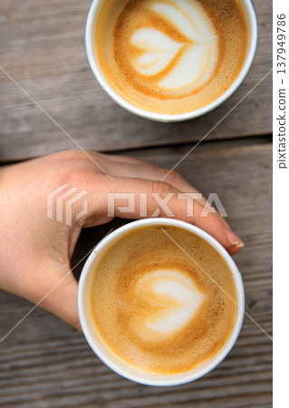 Closeup of a hand holding a paper coffee cup with heart-shaped latte art beside another cup on a rustic wooden table. Closeup of a hand holding a paper coffee cup with heart-shaped latte art beside another cup on a rustic wooden table. 137949786