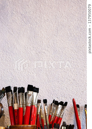 Closeup of paintbrushes with red handles, shiny metal ferrules and dense black tufts, neatly arranged in glass jars and clay jugs, set against a textured wall. 137950079