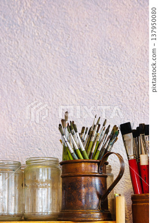 Closeup of assorted paintbrushes stored in a vintage copper pitcher and empty glass jars against a textured wall Closeup of assorted paintbrushes stored in a vintage copper pitcher and empty glass jars against a textured wall 137950080