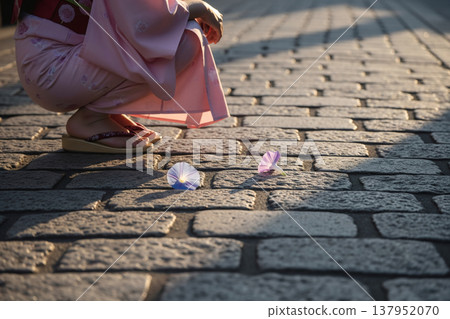 黃昏時分，一名身穿浴衣的女子蹲在鵝卵石街道上，周圍散落著牛花。 137952070