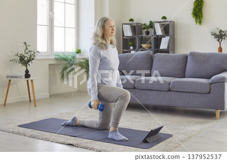 Senior woman doing sport exercise with dumbbells on yoga mat with tablet during online training. Senior woman doing sport exercise with dumbbells on yoga mat with tablet during online training. 137952537
