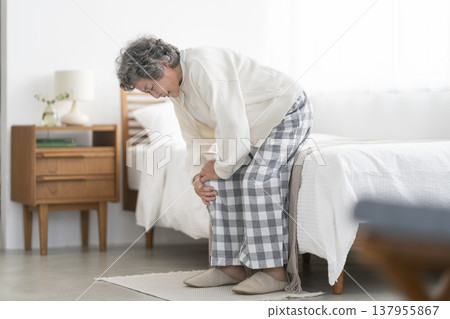 An 80-year-old woman holding her knees in front of the bed. 137955867
