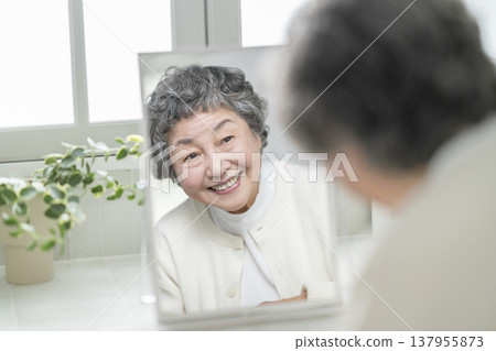 An 80-year-old woman smiles while looking in a mirror. Skincare & Beauty Image 137955873
