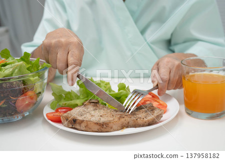 Asian elderly woman patient eating pork chop stake and vegetable salad for healthy food in hospital. Asian elderly woman patient eating pork chop stake and vegetable salad for healthy food in hospital. 137958182