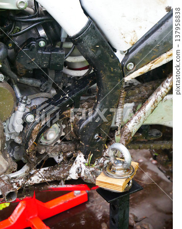 Dirty motorcycle engine and drive chain covered in mud. Muddy motocross bike frame resting on metal stand. Maintenance of dirt bike after offroad race. Extreme sport vehicle repair concept. Dirty motorcycle engine and drive chain covered in mud. Muddy motocross bike frame resting on metal stand. Maintenance of dirt bike after offroad race. Extreme sport vehicle repair concept. 137958838
