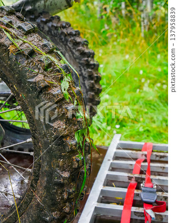 Muddy motorcycle tire on a metal ramp trailer. Extreme sport preparation after riding in nature. Off-road motorbike wheel with tread covered in dirt and grass during outdoor trip. Muddy motorcycle tire on a metal ramp trailer. Extreme sport preparation after riding in nature. Off-road motorbike wheel with tread covered in dirt and grass during outdoor trip. 137958839