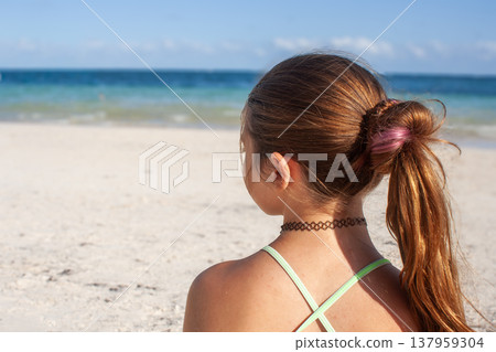 Close-up photo from behind of a teenager sitting on a white sand beach in Tulum, Mexico, gazing at the beautiful turquoise ocean under a bright blue sky. Close-up photo from behind of a teenager sitting on a white sand beach in Tulum, Mexico, gazing at the beautiful turquoise ocean under a bright blue sky. 137959304
