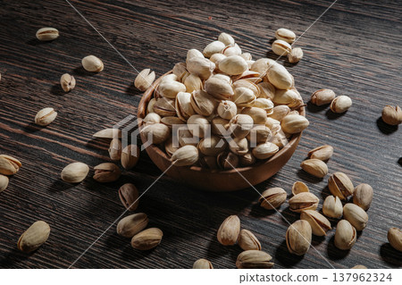 Wooden bowl filled with pistachios on dark wooden table, tasty salted nuts scattered around, healthy snack and natural food concept. Wooden bowl filled with pistachios on dark wooden table, tasty salted nuts scattered around, healthy snack and natural food concept. 137962324