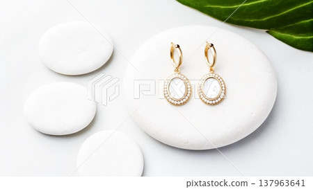 Elegant gold earrings with white stones displayed on smooth white pebbles and green leaf 137963641