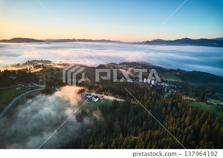 Aerial view of misty morning in mountains, with small village nestled among trees. Fog gently blankets landscape, creating serene and picturesque scene, as sun begins to rise in the background. Aerial view of misty morning in mountains, with small village nestled among trees. Fog gently blankets landscape, creating serene and picturesque scene, as sun begins to rise in the background. 137964192