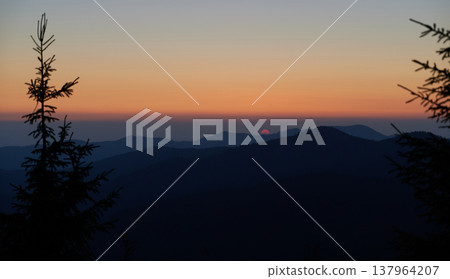 Sun sets behind distant mountains, casting soft orange and pink glow across sky. Silhouetted pine trees frame foreground, while layers of dark hills create serene and tranquil scene. Sun sets behind distant mountains, casting soft orange and pink glow across sky. Silhouetted pine trees frame foreground, while layers of dark hills create serene and tranquil scene. 137964207