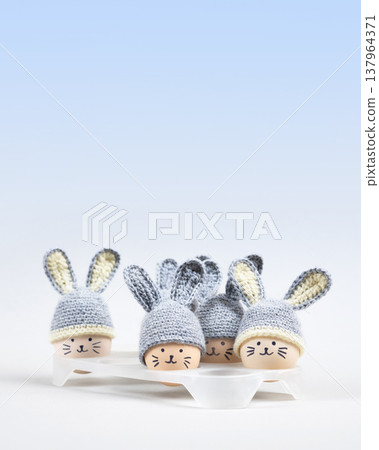Five Easter eggs with funny faces and gray crocheted hats with bunny ears in a white egg stand on a gray blue gradient background. Happy Easter concept. Greeting card. 137964371