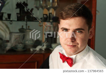 Portrait of a young man in a white shirt and red bow tie. Handsome male sitting in an interior setting with a blurred vintage cabinet background. Man 20s years old Portrait of a young man in a white shirt and red bow tie. Handsome male sitting in an interior setting with a blurred vintage cabinet background. Man 20s years old 137964565