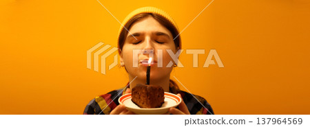 Young woman with closed eyes blowing out a candle on a chocolate cake. Birthday celebration and making a wish concept on a yellow background. Happy Birthday banner 137964569