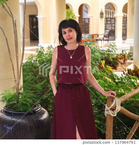 Portrait of a brunette in a burgundy dress standing in a lobby near green plants 137964761