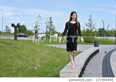 Woman in black dress walking barefoot on pavement in a park on a sunny day. Woman in black dress walking barefoot on pavement in a park on a sunny day. 137966498