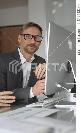 Business professional analyzing growing financial data on computer monitor with colleague, pointing at screen while discussing market trends and economy strategy in modern office, vertical view 137968036