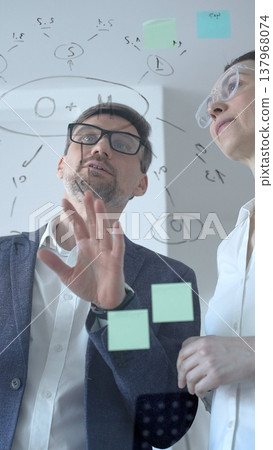 Business professionals brainstorming analyzing data and developing new strategies on a transparent whiteboard with handwritten diagrams and digital tablet 137968074