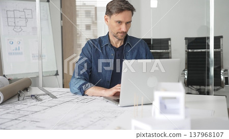 Male architect wearing denim shirt working on laptop in modern office, analyzing blueprints and architectural plans on desk, designing new building construction project Male architect wearing denim shirt working on laptop in modern office, analyzing blueprints and architectural plans on desk, designing new building construction project 137968107