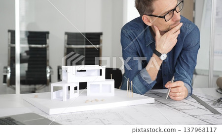 Male architect thinking while looking at a scale model of a modern house, developing architectural plans and blueprints for a new residential project in an office setting Male architect thinking while looking at a scale model of a modern house, developing architectural plans and blueprints for a new residential project in an office setting 137968117