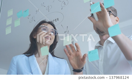 Business colleagues collaborating on strategy, brainstorming ideas for success, writing on a transparent glass board with sticky notes in a modern office Business colleagues collaborating on strategy, brainstorming ideas for success, writing on a transparent glass board with sticky notes in a modern office 137968124