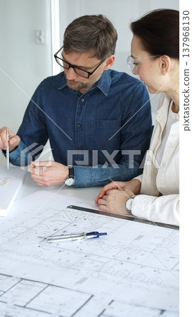 Architects and engineers collaborate over detailed blueprints and a house model, using compass and drafting tools to plan modern residential development with focused teamwork Architects and engineers collaborate over detailed blueprints and a house model, using compass and drafting tools to plan modern residential development with focused teamwork 137968130