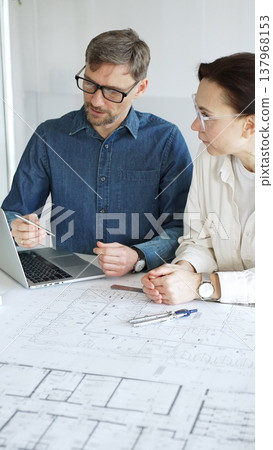 Architects and engineers collaborate over detailed blueprints and a house model, using compass and drafting tools to plan modern residential development with focused teamwork Architects and engineers collaborate over detailed blueprints and a house model, using compass and drafting tools to plan modern residential development with focused teamwork 137968153