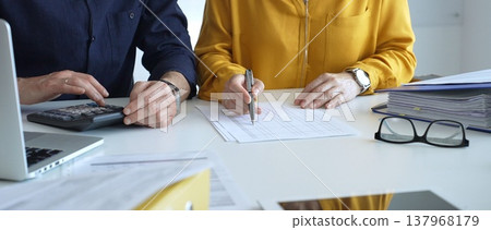 Two accountants collaborating in a well lit office, analyzing financial data and reviewing statements with a calculator and pen. Audit and taxes theme in business 137968179