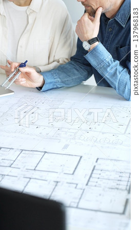 Engineering professionals collaborating on an architectural blueprint, using a compass to design a modern building, planning construction with a house model and laptop Engineering professionals collaborating on an architectural blueprint, using a compass to design a modern building, planning construction with a house model and laptop 137968183