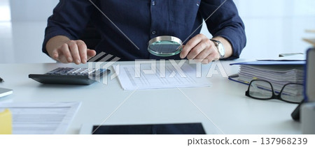 Male financial analyst reviewing paperwork, using calculator and magnifying glass while examining corporate budget reports in modern office. Audit and taxes, business concept 137968239