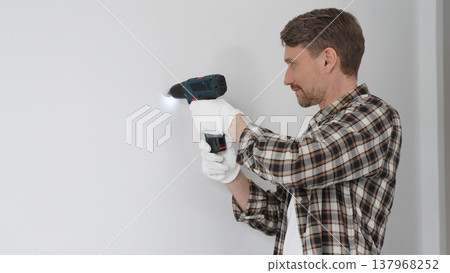 Man construction worker in checked shirt and safety gloves is screwing a screw into the wall using a cordless screwdriver, carrying out a careful home renovation. Horizontal portrait view 137968252