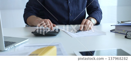 Businessman using a calculator and analyzing financial documents while preparing paperwork at an office desk. Audit and taxes, business concept 137968282