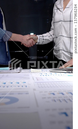 Professionals shaking hands at night over a desk full of financial charts and a tablet, confirming a successful business agreement after a late meeting Professionals shaking hands at night over a desk full of financial charts and a tablet, confirming a successful business agreement after a late meeting 137968284