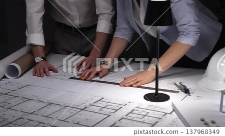 Collaborative team working on an engineering project, reviewing floor plans with a lamp illuminating the architectural drawings. Business and architect people concept Collaborative team working on an engineering project, reviewing floor plans with a lamp illuminating the architectural drawings. Business and architect people concept 137968349