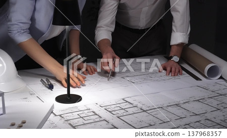 Collaborative team working on an engineering project, reviewing floor plans with a lamp illuminating the architectural drawings. Business and architect people concept Collaborative team working on an engineering project, reviewing floor plans with a lamp illuminating the architectural drawings. Business and architect people concept 137968357