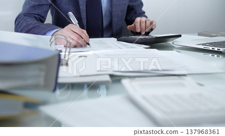 Businessman or accountant wearing blue suit is working with laptop, calculator and documents in office. Low lighting, close-up view. Business people, audit concept Businessman or accountant wearing blue suit is working with laptop, calculator and documents in office. Low lighting, close-up view. Business people, audit concept 137968361