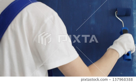 Painter applying blue paint with roller on white wall, showing precise technique and transforming interior space with smooth, even strokes 137968388