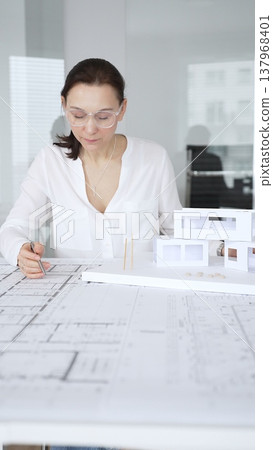 Female architect in eyeglasses concentrating on a detailed building model and blueprints, carefully planning a modern architectural design in a professional office 137968401