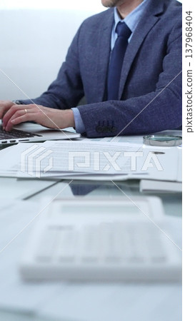 Businessman wearing blue suit is working with laptop, calculator and documents at the glass table in office set. Low lighting, close-up vertical view. Business people and audit 137968404