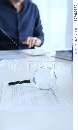 Magnifying glass highlighting a financial report with a businessman working on a laptop in the blurred background. Audit and taxes in business Magnifying glass highlighting a financial report with a businessman working on a laptop in the blurred background. Audit and taxes in business 137968422