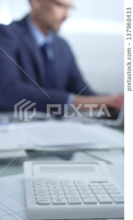 Calculator, pen, laptop and documents are lying on accountant's desk. Businessman wearing blue suit is working with documents on the background. Vertical view. Business and audit concept Calculator, pen, laptop and documents are lying on accountant's desk. Businessman wearing blue suit is working with documents on the background. Vertical view. Business and audit concept 137968433