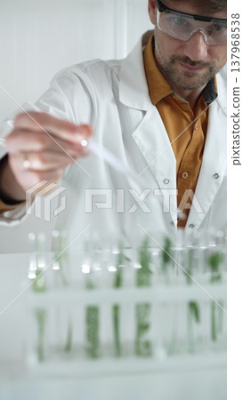 A male science researcher, wearing a lab coat and protective glasses, drips liquid from a pipette into a test tube containing a green plant. His face is focused, captured in a vertical portrait 137968538