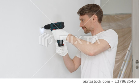 Professional male worker wearing white t-shirt and protective gloves, is drilling hole in a wall with cordless drill during home renovation work. Horizontal portrait view Professional male worker wearing white t-shirt and protective gloves, is drilling hole in a wall with cordless drill during home renovation work. Horizontal portrait view 137968540