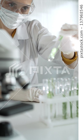 Woman scientist wearing lab coat, white gloves, face mask and protective glasses, is holding a test tube with plants inside near microscope in laboratory, vertical portrait view. Science and medicine 137968546