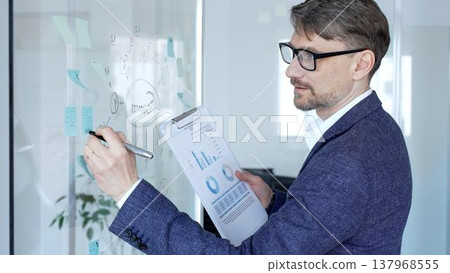 Mature businessman in glasses actively analyzing financial data, developing business strategy, brainstorming ideas, and planning for success on a modern glass whiteboard in the office setting Mature businessman in glasses actively analyzing financial data, developing business strategy, brainstorming ideas, and planning for success on a modern glass whiteboard in the office setting 137968555