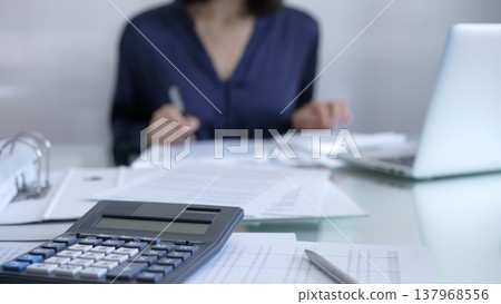 Black calculator, pen and documents are lying on accountant's desk. Businesswoman wearing blue dress is working with documents on the background. Business and audit concept Black calculator, pen and documents are lying on accountant's desk. Businesswoman wearing blue dress is working with documents on the background. Business and audit concept 137968556
