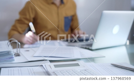 Magnifying glass, calculator, pen and documents are lying on accountant's desk. Businessman or accountant, wearing yellow shirt, is working with documents on the background. Business people, audit Magnifying glass, calculator, pen and documents are lying on accountant's desk. Businessman or accountant, wearing yellow shirt, is working with documents on the background. Business people, audit 137968569